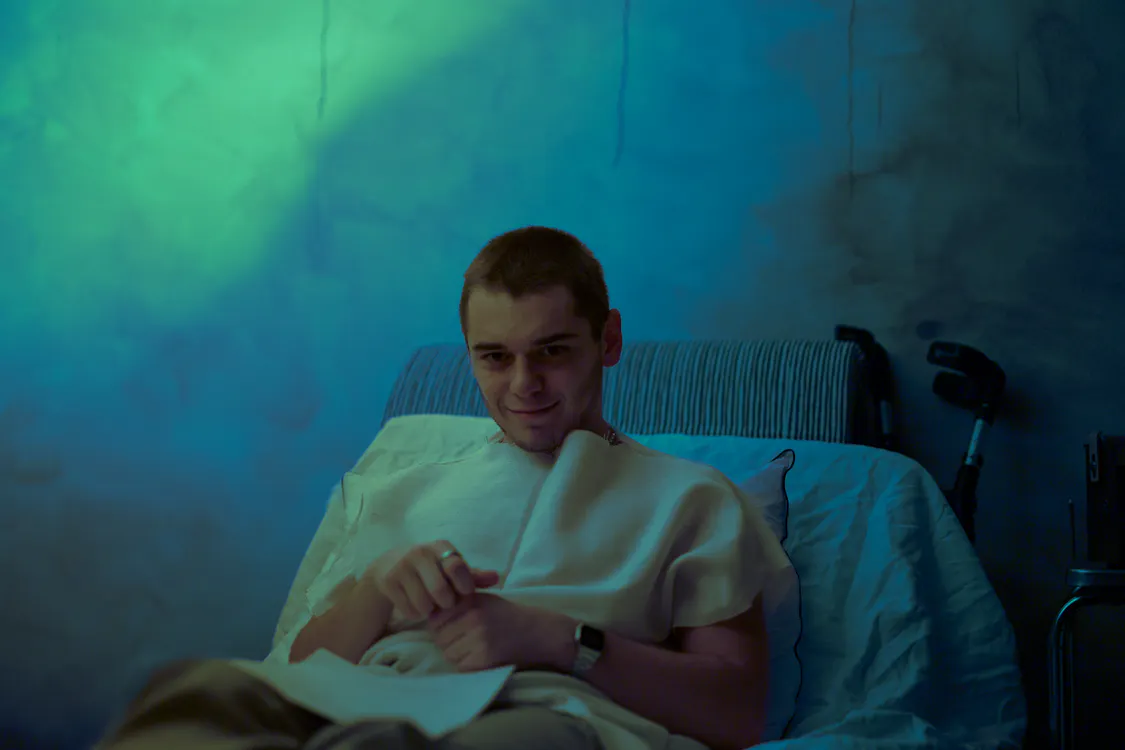 A smiling man with short hair relaxing in a hospital bed indoors.
